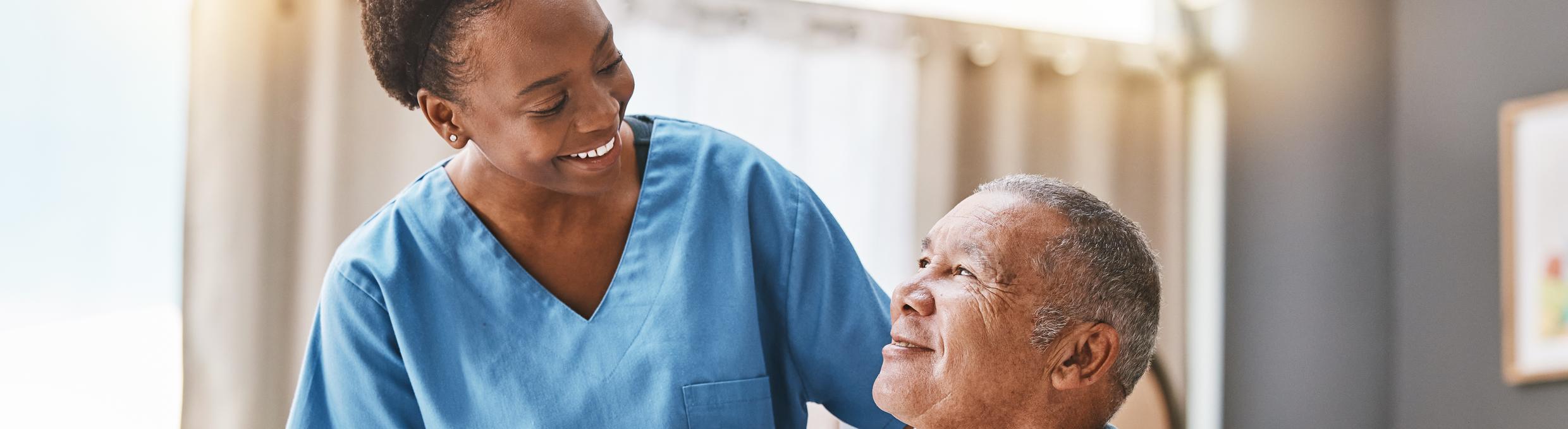A smiling caregiver with her hand on the shoulder of an older man in a wheelchair