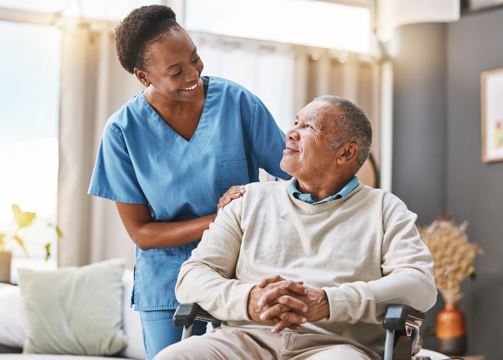 A caregiver with her hand on the shoulder of an older man in a wheelchair, both smiling